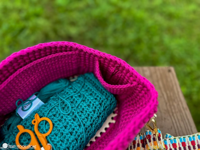 May include: A close-up of a magenta crochet bag filled with teal yarn, a crocheted teal item, and orange scissors. The bag has a textured appearance and a colorful strap. The background is blurred green.