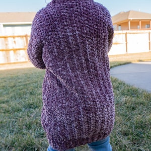 May include: A purple crocheted cardigan with a textured stitch pattern. The cardigan is worn by a child standing in a grassy area.