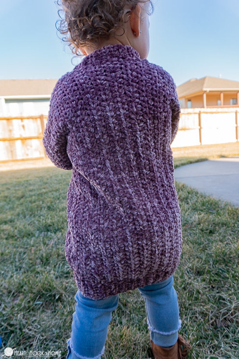 May include: A purple crocheted cardigan with a textured stitch pattern. The cardigan is worn by a child standing in a grassy area.
