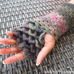 May include: A crocheted wrist warmer with a dragon scale pattern in shades of grey, green, and pink. The wrist warmer is made with a textured stitch and has a slightly ruffled edge.