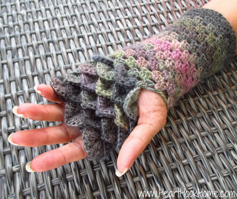 May include: A crocheted wrist warmer with a dragon scale pattern in shades of grey, green, and pink. The wrist warmer is made with a textured stitch and has a slightly ruffled edge.