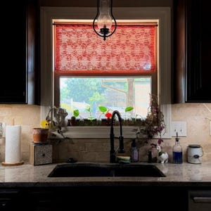 May include: A red crocheted valance hangs over a kitchen window above a black sink. The kitchen features dark brown cabinets, a granite countertop, a paper towel holder, a green pitcher, a small potted plant, and a white hanging fruit hammock.