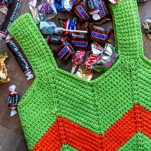 May include: A handmade green and orange crochet tote bag filled with various wrapped candies. The bag features a chevron design and is surrounded by candy wrappers, including Snickers, Twix, and Tootsie Roll.