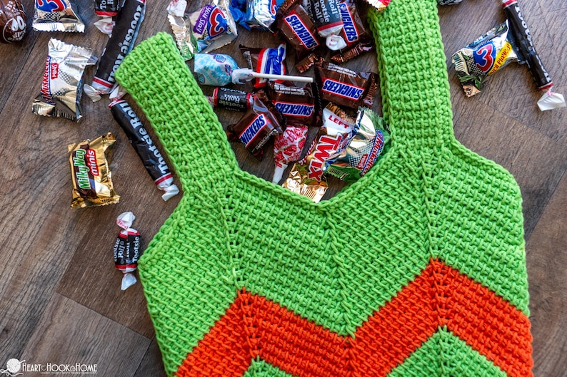 May include: A handmade green and orange crochet tote bag filled with various wrapped candies. The bag features a chevron design and is surrounded by candy wrappers, including Snickers, Twix, and Tootsie Roll.