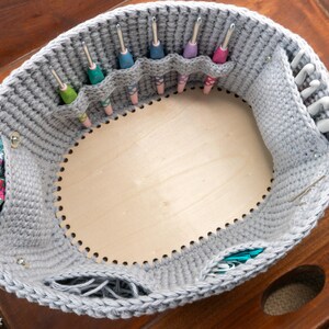 May include: A gray crochet basket with a wooden base, designed to hold crochet hooks. The basket has several pockets for organizing hooks of various sizes and colors. A decorative fabric is attached to the side.