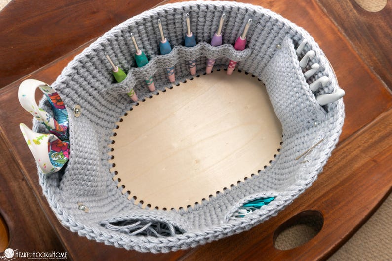May include: A gray crochet basket with a wooden base, designed to hold crochet hooks. The basket has several pockets for organizing hooks of various sizes and colors. A decorative fabric is attached to the side.