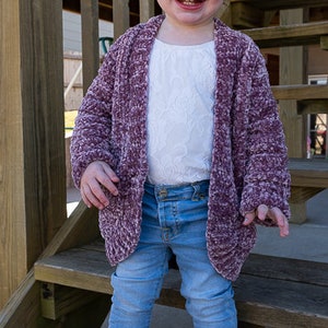 May include: A young girl wearing a purple fuzzy cardigan over a white lace top, blue jeans, and brown suede boots with fringe. She is standing on wooden steps and has a white bow in her hair.