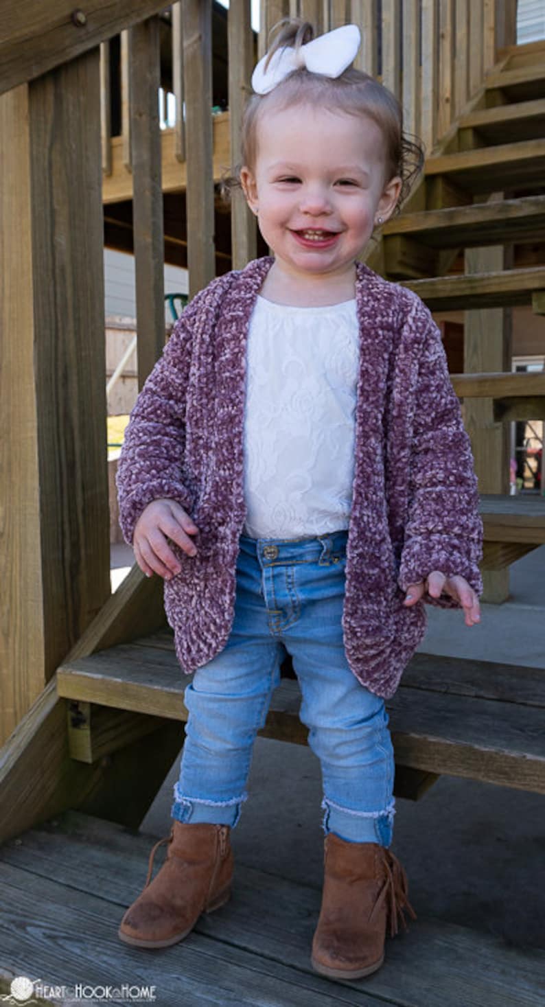 May include: A young girl wearing a purple fuzzy cardigan over a white lace top, blue jeans, and brown suede boots with fringe. She is standing on wooden steps and has a white bow in her hair.