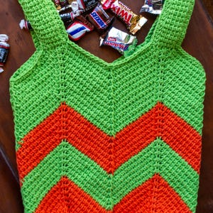 May include: A handmade, green and orange crochet tote bag with a chevron pattern. The bag is filled with various candies, including Snickers, Twix, and Tootsie Rolls. The bag has shoulder straps and is made of a textured yarn.