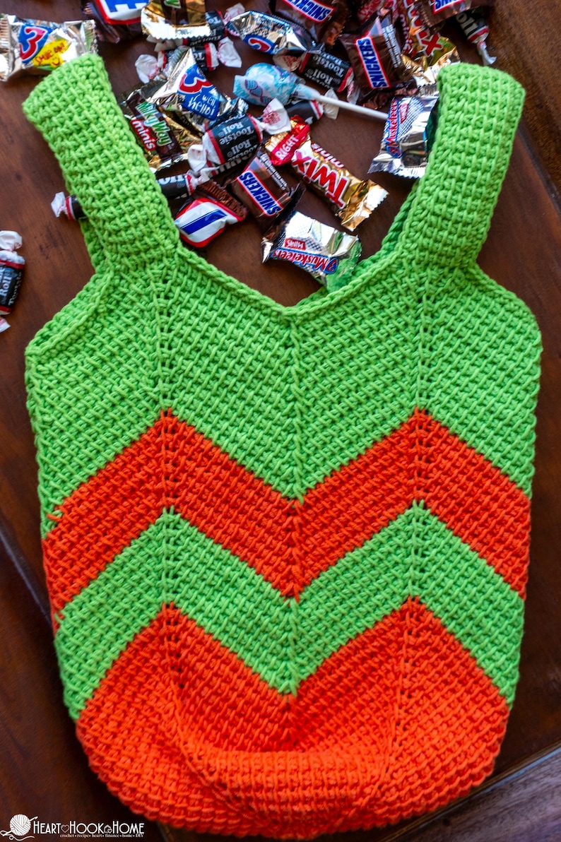 May include: A handmade, green and orange crochet tote bag with a chevron pattern. The bag is filled with various candies, including Snickers, Twix, and Tootsie Rolls. The bag has shoulder straps and is made of a textured yarn.