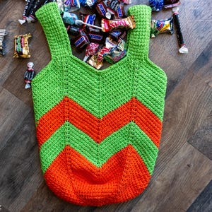 May include: A handmade crochet tote bag in green and orange chevron pattern. The bag has two straps and is filled with various candies. The bag is sitting on a wooden surface.