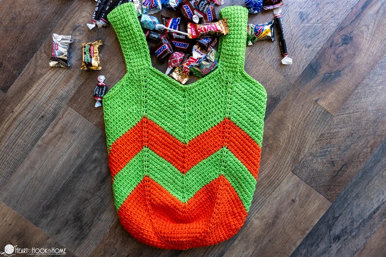 May include: A handmade crochet tote bag in green and orange chevron pattern. The bag has two straps and is filled with various candies. The bag is sitting on a wooden surface.