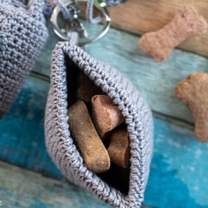 May include: A gray crocheted dog treat pouch with a drawstring closure and a metal key ring. The pouch is filled with brown dog biscuits.