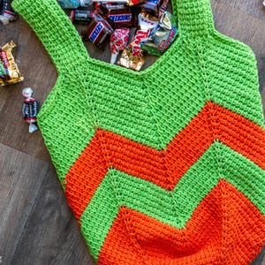 May include: A handmade crochet tote bag in green and orange chevron pattern, filled with various wrapped candies. The bag has two handles and is lying on a wooden surface.