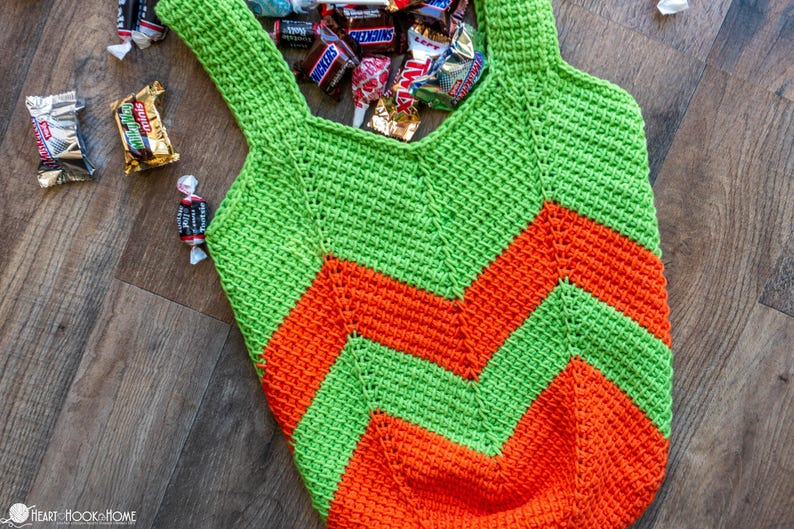 May include: A handmade crochet tote bag in green and orange chevron pattern, filled with various wrapped candies. The bag has two handles and is lying on a wooden surface.