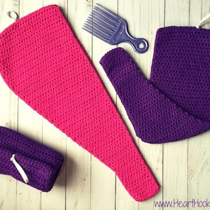 Pode incluir: Uma disposição plana de artigos de crochê feitos à mão em cores vibrantes. Uma toalha de crochê grande, rosa choque, com um laço para pendurar, uma toalha de crochê roxa e um pequeno embrulho de crochê roxo. Um pente roxo também está presente.
