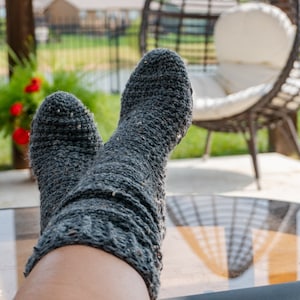 Peut inclure: Une paire de chaussettes en tricot gris avec un motif texturé. Les chaussettes sont portées par une personne se relaxant sur une terrasse avec un fauteuil en osier en arrière-plan.