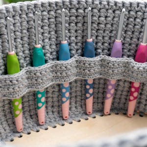 May include: A gray crocheted basket holds six crochet hooks. The hooks have colorful handles in green, teal, blue, purple, and pink, with pink and white polka dot accents. The basket has a built-in holder for the hooks.
