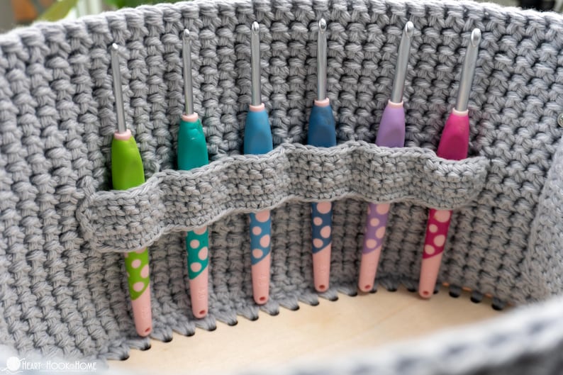May include: A gray crocheted basket holds six crochet hooks. The hooks have colorful handles in green, teal, blue, purple, and pink, with pink and white polka dot accents. The basket has a built-in holder for the hooks.