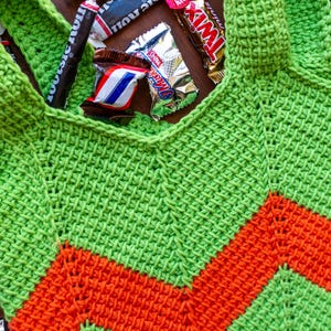 May include: A handmade green and orange crochet tote bag with a chevron design. The bag is filled with various wrapped candies, including Tootsie Rolls, Twix, and Snickers bars. The bag has two shoulder straps.