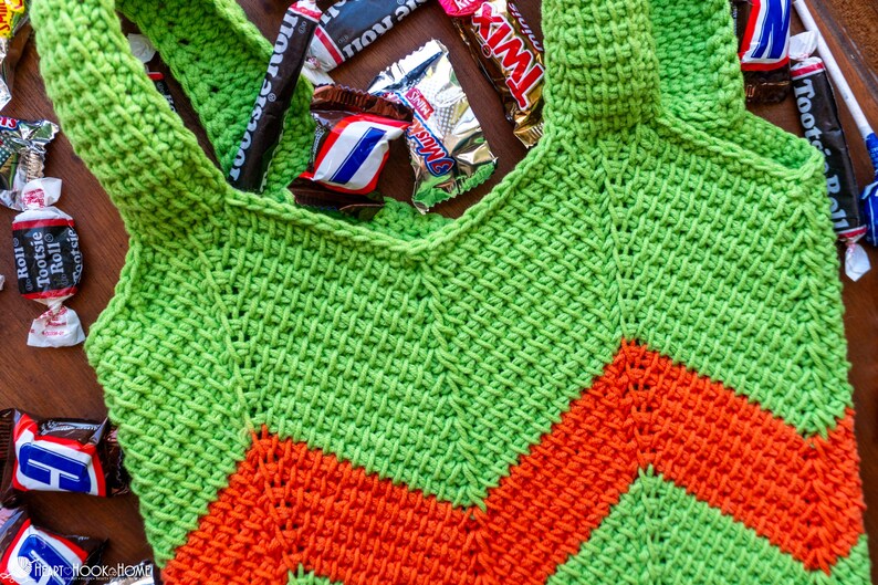 May include: A handmade green and orange crochet tote bag with a chevron design. The bag is filled with various wrapped candies, including Tootsie Rolls, Twix, and Snickers bars. The bag has two shoulder straps.