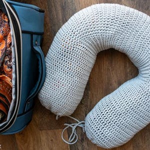 Puede incluir: Una almohada de viaje de crochet gris claro descansa sobre una superficie de madera junto a una bolsa de viaje azul. La bolsa est&aacute; abierta, revelando ropa y un cintur&oacute;n enrollado. La almohada tiene forma de U, dise&ntilde;ada para soporte del cuello.