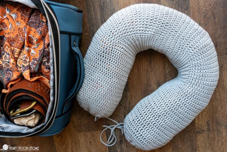 Puede incluir: Una almohada de viaje de crochet gris claro descansa sobre una superficie de madera junto a una bolsa de viaje azul. La bolsa est&aacute; abierta, revelando ropa y un cintur&oacute;n enrollado. La almohada tiene forma de U, dise&ntilde;ada para soporte del cuello.
