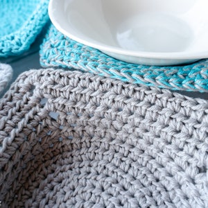 May include: Close-up of a gray crocheted coaster with a white bowl on top. The coaster is made with a simple stitch pattern and has a slightly textured surface.