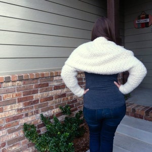 May include: A white crocheted shrug with long sleeves. The shrug is worn over a gray shirt and blue jeans.