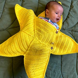 May include: A baby wrapped in a bright yellow, star-shaped crocheted blanket. The blanket has a button closure and a star-shaped button. The baby is wearing a striped shirt and is lying on a green blanket.