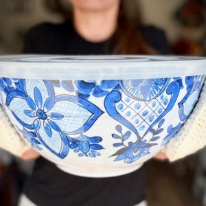 May include: A large white mixing bowl with a blue floral pattern and a translucent plastic lid, held by two cream-colored crocheted bowl cozies. The bowl features intricate blue petal and lattice designs across its exterior surface.