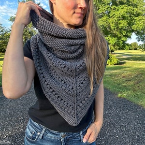 May include: A gray triangular shawl with a lacy pattern and a textured knit. The shawl is worn by a person with long brown hair.