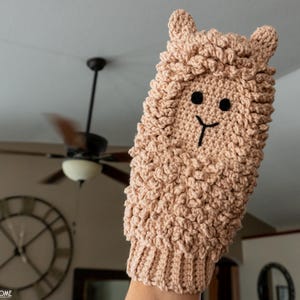 May include: A tan crochet llama hand puppet with a textured, fluffy body. The puppet has black eyes and a black smile. The ears are small and upright. The cuff is ribbed. The puppet is held up against a blurred background.