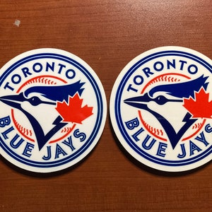 Peut inclure: Deux autocollants circulaires avec le logo des Blue Jays de Toronto. Le logo comprend un geai bleu, une feuille d'érable rouge et les mots "TORONTO" et "BLUE JAYS" en bleu et blanc. Sur une surface marron.