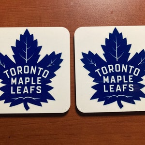 Peut inclure: Deux sous-verres carrés blancs avec un logo bleu des Maple Leafs de Toronto. Le logo est une feuille d'érable stylisée avec les mots "Toronto Maple Leafs" en texte blanc.