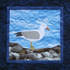 May include: A white seagull with a yellow beak and legs stands on a rocky shore. The seagull is perched on a bed of grey and brown rocks. The background is a blue and white sky with clouds.