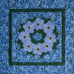 May include: A blue and green floral quilt block with a wreath of forget-me-not flowers in the center.