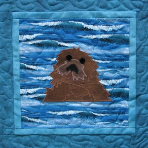 May include: A brown and white sea otter with black eyes is appliqued on a blue and white fabric background. The fabric has a wave pattern.