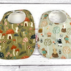 May include: Two baby bibs with woodland animal and mushroom prints. The bib on the left has a green background with mushrooms, leaves, and mice. The bib on the right has a light green background with trees, animals, and a campfire.