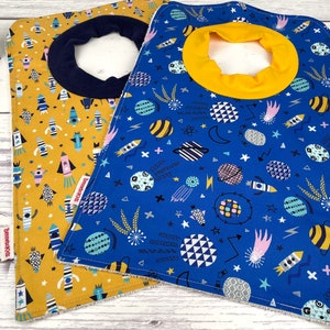 May include: Two baby bibs with space-themed patterns. One bib is mustard yellow with rocket ships, the other is blue with planets, stars, and rockets. Each bib has a contrasting colored neck opening: navy blue and yellow.
