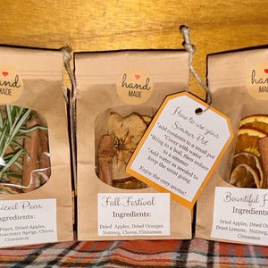 Op de afbeelding: Drie bruine papieren zakken met gedroogde vruchten en kruiden voor een sudderpot. Elke zak heeft een doorzichtig venster, een handgemaakte sticker en een etiket met de ingrediënten voor Spiced Pear, Fall Festival en Bountiful Harvest, plus een instructielabel.
