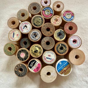 May include: A collection of vintage wooden sewing thread spools arranged in a cluster. Each spool features a unique circular paper label on the top, displaying various brand names like Clark's, Belding, and Talon, along with thread color numbers.