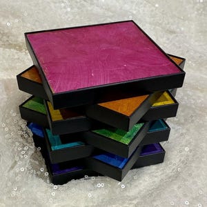 May include: A stack of square coasters with black frames and textured, multi-colored surfaces. The top coaster is magenta, followed by layers in orange, yellow, green, blue, and purple. Each coaster features a distinct, hand-painted appearance.