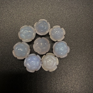 May include: Eight white, opaque, round, decorative beads with a floral design. The beads are arranged in a circle on a dark gray surface.
