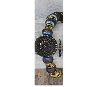 May include: A bracelet with blue and gold beads and a large, round, black metal charm with a floral design. The charm has a raised, textured border. The bracelet is labeled "royal crown".
