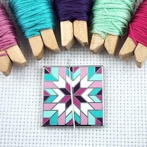 May include: A silver metal pin with a colorful geometric design. The pin features a starburst pattern in shades of pink, teal, and purple. A silver needle is attached to the pin.