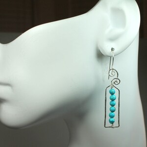 May include: Silver dangle earrings with a turquoise bead design. The earrings feature a rectangular frame with a series of turquoise beads hanging from it. The earrings are attached to a silver hook.