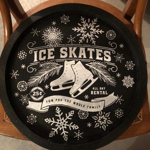 May include: Black and white round sign with the text "ICE SKATES" in a white banner. The sign also includes a pair of white ice skates, snowflakes, and the text "25c", "ALL DAY RENTAL", and "FUN FOR THE WHOLE FAMILY".
