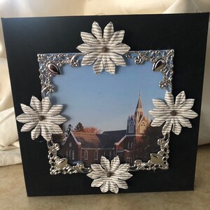 Puede incluir: Un marco de fotos negro con un borde decorativo plateado y cuatro flores de papel rayadas blancas y grises. El marco contiene una foto de un edificio de iglesia con un campanario.