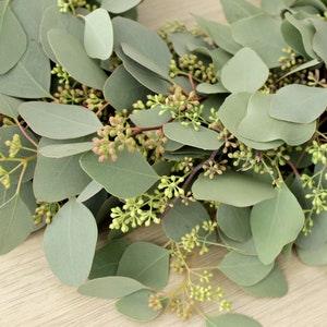May include: A close-up of a eucalyptus garland with green leaves and small green buds. The garland is on a light wood surface.